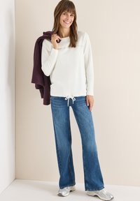 Witte lange mouwen top met een trekkoord aan de onderkant, gecombineerd met blauwe flared jeans. Een paarse jas over de arm gedrapeerd, en zilveren sneakers maken de look compleet.
