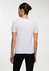 Donna con acconciatura a ciuffo intrecciato, che indossa una semplice t-shirt bianca a maniche corte e pantaloni neri, rivolta di spalle su uno sfondo bianco.