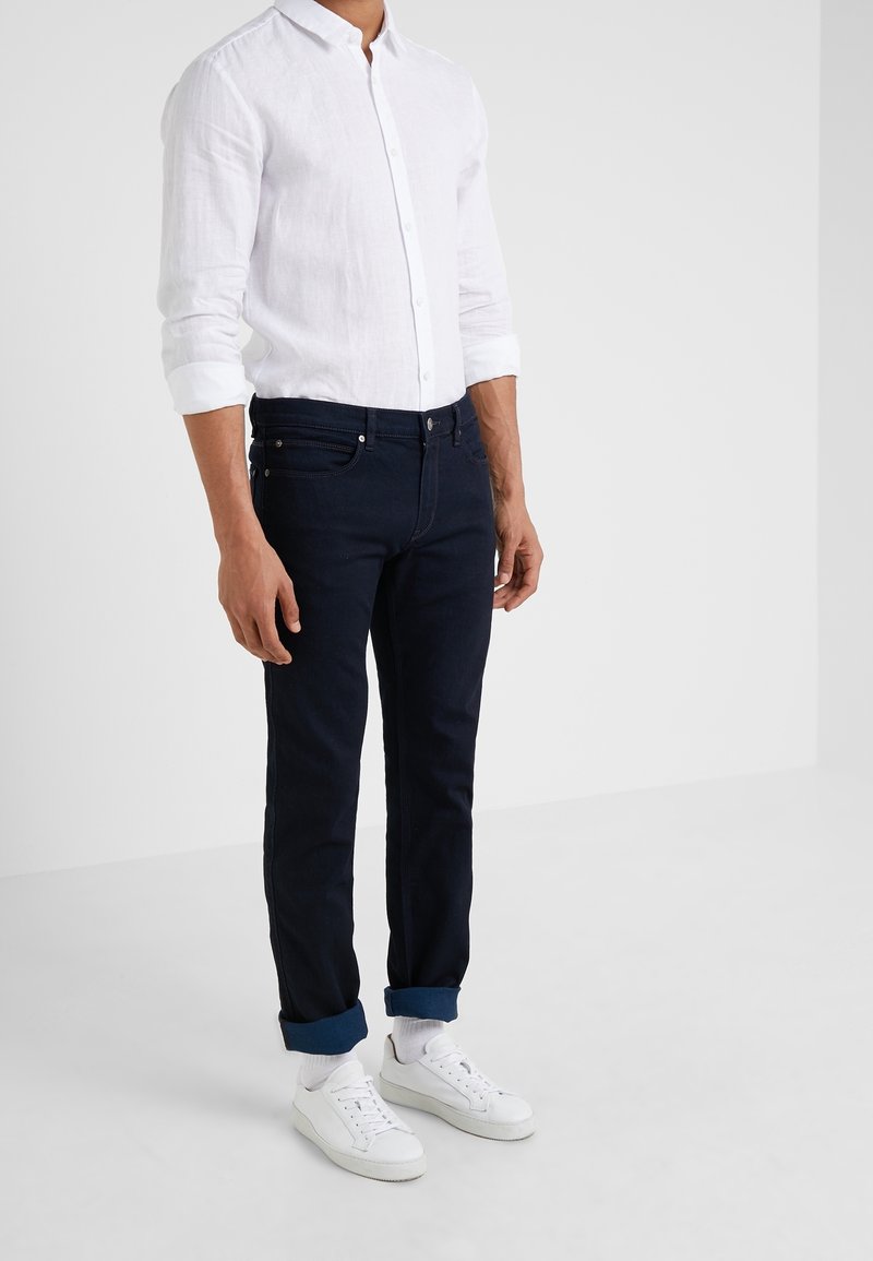 Homme portant une chemise blanche boutonnée aux manches retroussées, un jean sombre avec revers, des chaussettes blanches et des baskets blanches, debout contre un mur uni.