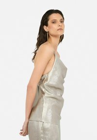 Top senza maniche in argento metallizzato con design drappeggiato; texture liscia. Il modello indossa orecchini di dichiarazione, capelli acconciati sciolti. Sfondo semplice.