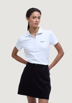 Kobieta z zaplecionymi włosami ma na sobie białą koszulkę polo Barbour i czarną spódnicę, stoi z rękami w kieszeniach spódnicy na jednolitym tle.