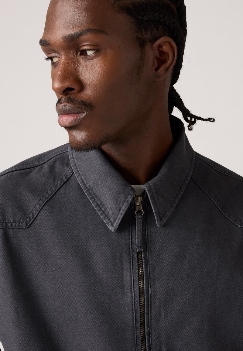 Dunkelgraue Zip-Jacke mit spitzem Kragen und sichtbaren Stichelementen. Sie verfügt über einen bronzefarbenen Reißverschluss und einen strukturierten Stoff.