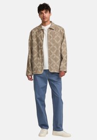 Beige patterned shirt jacket with a collar, button closure, and single chest pocket, paired with light blue trousers and white sneakers.
