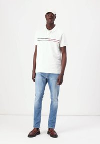 Camisa polo blanca con detalles en franjas rojas y marinas, jeans azul claros, mocasines marrones y una gorra beige. Diseño minimalista y estilo casual.