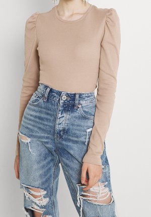 Nærbillede af en person iført en beige langærmet top med puffede skuldre og højtaljede, kraftigt revnede blå jeans.