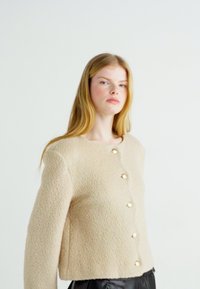 Beige lyhythihainen takki tekstitetulla pinnalla, jossa on viisi kultasävyistä nappia edessä ja pyöreä kaula-aukko. Mustat housut on pukeutunut alle.