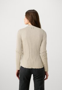 Beige ribbet langermet topp med krage, subtil glans og buet hem. Kombinert med sorte jeans, sett fra baksiden.