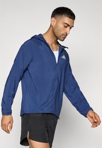 Ελαφρύ μπλε ναυτικό μπουφάν με κουκούλα, φερμουάρ κλεισίματος και τρεις λευκές διαγώνιες ρίγες στο πάνω αριστερό στήθος. Ομαλή υφή και αθλητικός σχεδιασμός.