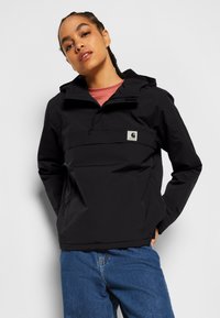 Giacca a pullover nera impermeabile con tasca frontale, cappuccio regolabile e patch con logo. Indossata con una maglietta rosa e jeans blu.