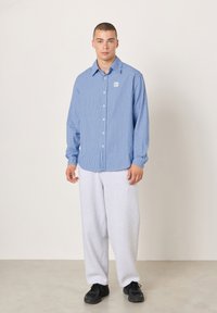 Čovjek stoji noseći plavu prugastu košulju s gumbima, svijetlosive široke hlače i crne tenisice, na jednostavnoj svijetloj pozadini.