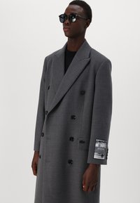 Cappotto grigio doppiopetto in tessuto strutturato con bottoni neri e una toppa sulla manica. Indossato con occhiali da sole neri e un top nero.