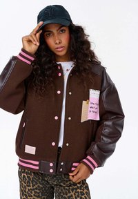 Młoda kobieta z kręconymi włosami, nosząca brązową kurtkę bomberkę z różowymi akcentami, spodnie w panterkę i czarną czapkę, poprawiająca czapkę jedną ręką.