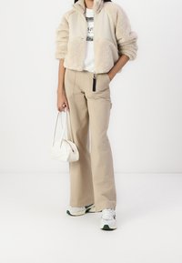 Beżowa, puszysta kurtka z zamkiem i kołnierzem, biała koszulka z grafiką, jasnobeżowe szerokie spodnie, biała torebka i szaro-zielone sneakersy.