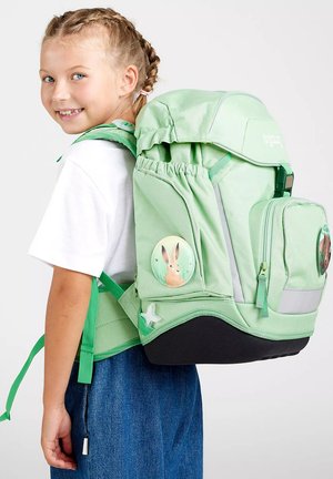 Junges Mädchen mit geflochtenem Haar lächelt und trägt einen großen hellgrünen Rucksack mit Hasen- und Bären-Aufnähern.