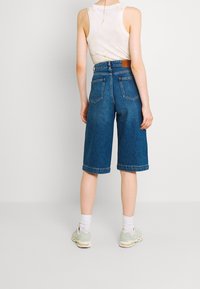 Denim bermuda shorts dengan pinggang tinggi, berwarna biru muda, tepi digulung, dan dua saku belakang. Dipadukan dengan tank top beige dan sneakers ringan.