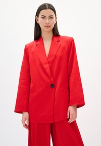 Roter oversized Blazer aus glattem Stoff, mit einem einzelnen Knopfverschluss, scharfen Revers und seitlichen Taschen. Lange, weite Ärmel.