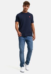 Camiseta de manga corta en color navy con un pequeño logo rojo en el pecho, combinada con jeans de color azul claro y zapatillas blancas. Diseño simple y casual.