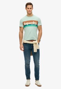 Man wearing mint green Superdry t-shirt, blue jeans, beige sweater tied at waist, white sneakers, and brown watch standing against white background.