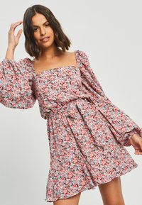 Cvetna obleka z kvadratnim izrezom, napihnjenimi rokavi in pasom. Tkanina prikazuje večbarvno cvetje na roza ozadju.