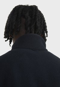 Veste en polaire bleu marine avec un col haut, texture douce et un petit détail en boucle sous le col, conçue pour la chaleur et le confort.