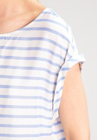 Blusa leggera di tessuto bianco con righe orizzontali blu, scollatura ampia e maniche corte arrotolate. Dettaglio delle cuciture visibile sul lato.