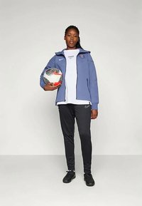 Blå sportsjakke med hette, hvit skjorte, svarte bukser og svarte fotballsko. Holder en mønstret fotball med røde detaljer.