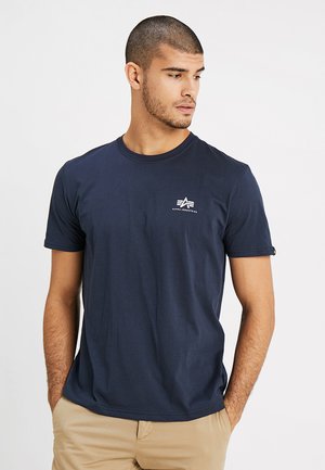Mann trägt ein dunkelblaues Alpha Industries T-Shirt und beige Hose, steht mit den Händen in den Taschen und schaut nach rechts.