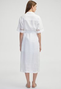 Witte linnen jurk met korte mouwen, een getailleerde taille en uitsparingen op de mouwen. Vloerlange stijl, minimale textuur.