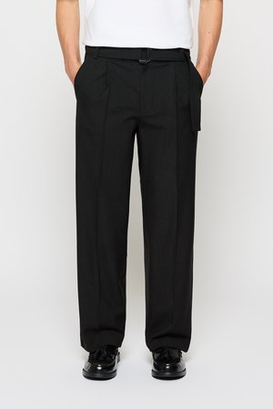 Man in zwarte streepbroek met riem en zwarte tassel instappers, staande met handen in de zakken tegen een witte achtergrond.
