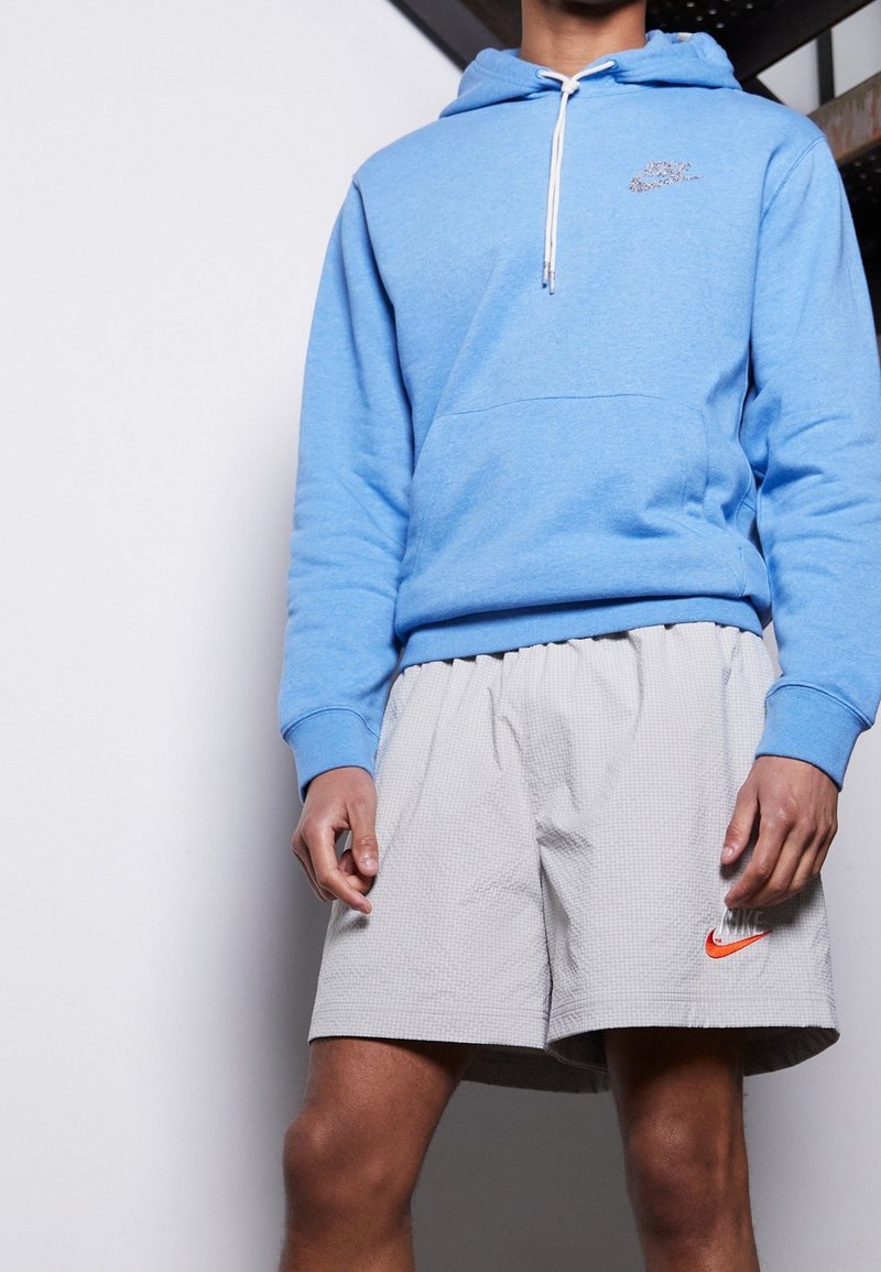 Ljusblå huvtröja med framficka och dragsko. Grå rutiga shorts med en orange Nike-logga på vänster lår.