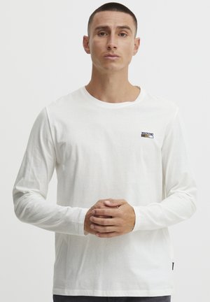 Camiseta blanca de manga larga, con un pequeño logo azul y amarillo de "WEEKEND" en el lado izquierdo del pecho, con cuello redondo y tejido texturizado.