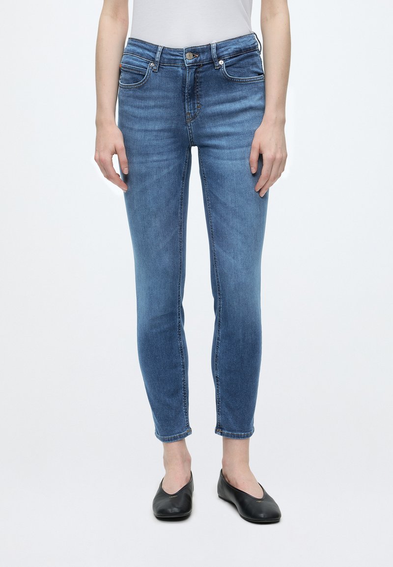 Personne portant un jean bleu ajusté et des chaussures plates noires, debout devant un fond blanc uni.