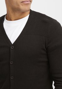 Mørkebrun cardigan med V-udskæring, ribbede skulderdetaljer og seks knaplukninger, lavet af et glat strikket stof.