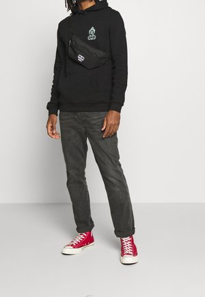 Sudadera negra con gráfico verde, jeans grises y zapatillas rojas de caña alta. Riñonera negra llevada en el pecho con cremallera y detalle de logo.