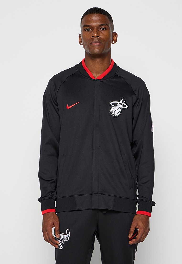 Giacca sportiva nera con dettagli rossi, dotata di zip frontale, polsini a coste e logo sul lato sinistro del petto. Tessuto liscio e leggero.