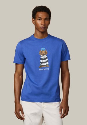 Homme portant un T-shirt bleu avec un motif d'un chien en chemise rayée et casquette, associé à un pantalon de couleur claire.