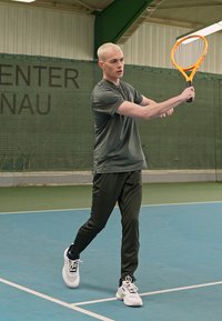 Sportler in einem grauen Sportshirt und dunklen Hosen, der einen orangefarbenen Tennisschläger hält und einen Schlag auf einem blauen Innenplatz ausführt.