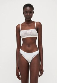 Štíhla žena s tmavou pokožkou, oblečená v bielom priesvitnom braletku s kvetinovým vzorom a bielych nohavičkách v štýle bikín s čipkovým okrajom, stojaca proti jednoliatej bielej pozadia.