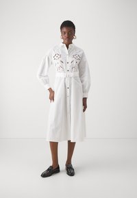 Abito bianco a camicia con bottoni sul davanti, vita affusolata e dettagli a intaglio floreale. Maniche lunghe con polsini arricciati, indossato con mocassini neri.
