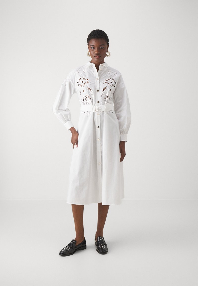 Abito bianco a camicia con bottoni sul davanti, vita affusolata e dettagli a intaglio floreale. Maniche lunghe con polsini arricciati, indossato con mocassini neri.