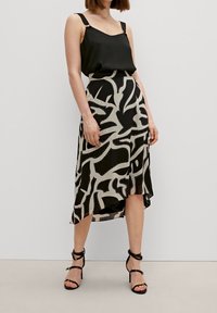 Zwarte mouwloze top gecombineerd met een beige rok met gedurfde zwarte abstracte patronen en spijkerdetails, een asymmetrische zoom, en strappy hakken.