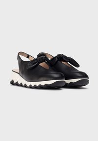 Zapatos de cuña de cuero negro con punta redonda y lados con cortes, que presentan un detalle de lazo en la parte delantera y una suela blanca con zig zag.
