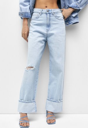 Hellblaue High-Waist Jeans mit weitem Bein, Knieschlitz und umgeschlagenen Säumen, getragen mit passenden blauen, offen zehigen Sandalen mit Riemchen.