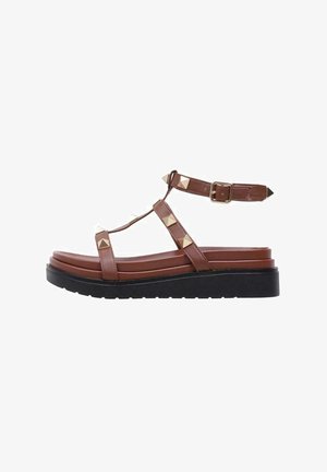 Sandalia de imitación cuero marrón con diseño de tira en T, tachuelas en forma de pirámide, correa de tobillo ajustable y suela de plataforma negra gruesa.