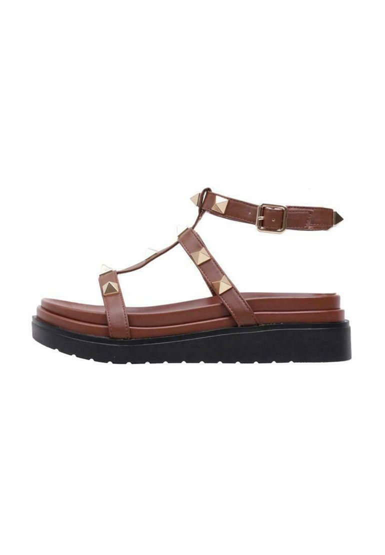 Sandale en faux cuir marron avec design en T, clous en forme de pyramide, sangle de cheville ajustable et semelle épaisse noire.