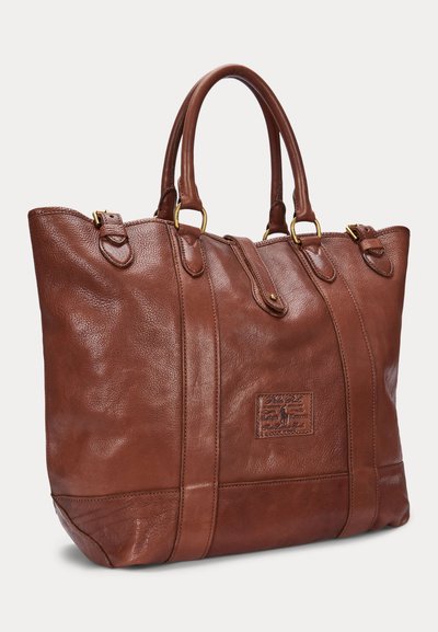 Sac fourre-tout en cuir marron avec double poignées, garnitures dorées et un patch de marque cousu sur le panneau avant.