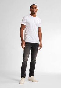 Camiseta blanca de algodón con un pequeño logo, combinada con jeans gris oscuro y zapatillas de color claro. Modelo posando sobre un fondo neutro.