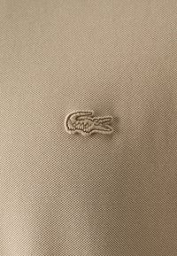 Tessuto beige con una texture che presenta un piccolo logo di coccodrillo ricamato nella stessa tonalità. Il design è minimal e semplice.