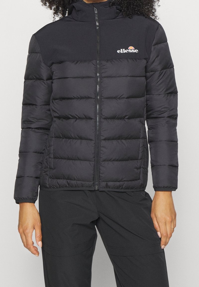 Chaqueta de plumas negra con cierre de cremallera, textura acolchada y puños elásticos. Presenta un logo en el lado izquierdo del pecho y una capucha.
