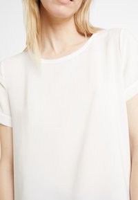 Blusa blanca transparente con escote redondo, mangas cortas y textura suave. Presenta un diseño simple, clásico y detalles minimalistas.